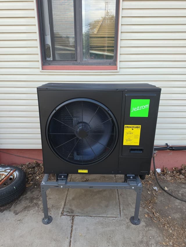 Black Jetson Air 3-ton heat pump mounted against cream vinyl siding on a concrete pad at a Pueblo, CO home.