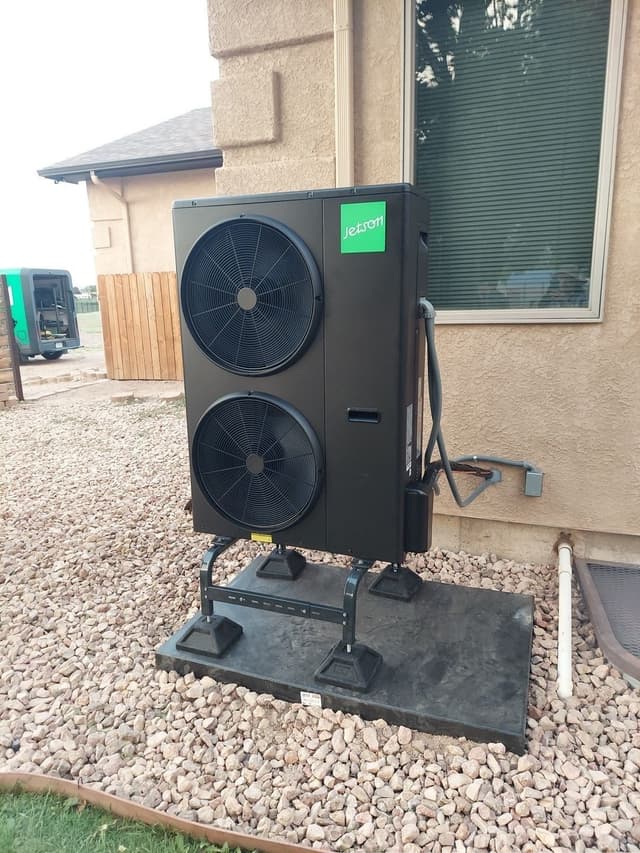 A black Jetson Air 4-ton heat pump installed along the tan stucco exterior wall of a Pueblo home, with conduit running neatly to a disconnect box and a gravel bed at the base.