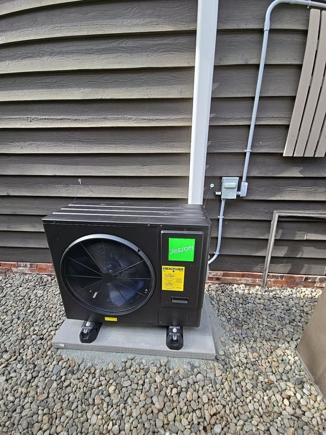 Black Jetson Air 3-ton central ducted heat pump condenser on a concrete pad in a gravel bed against dark grey lap siding of a home in Broadmoor, Richmond, BC.