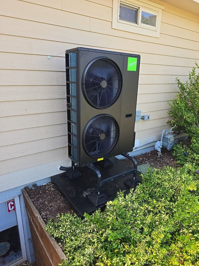 Jetson Air dual-fan 4-ton heat pump condenser on a black stand against tan lap siding, with shrubs on either side and a gas meter visible to the right.