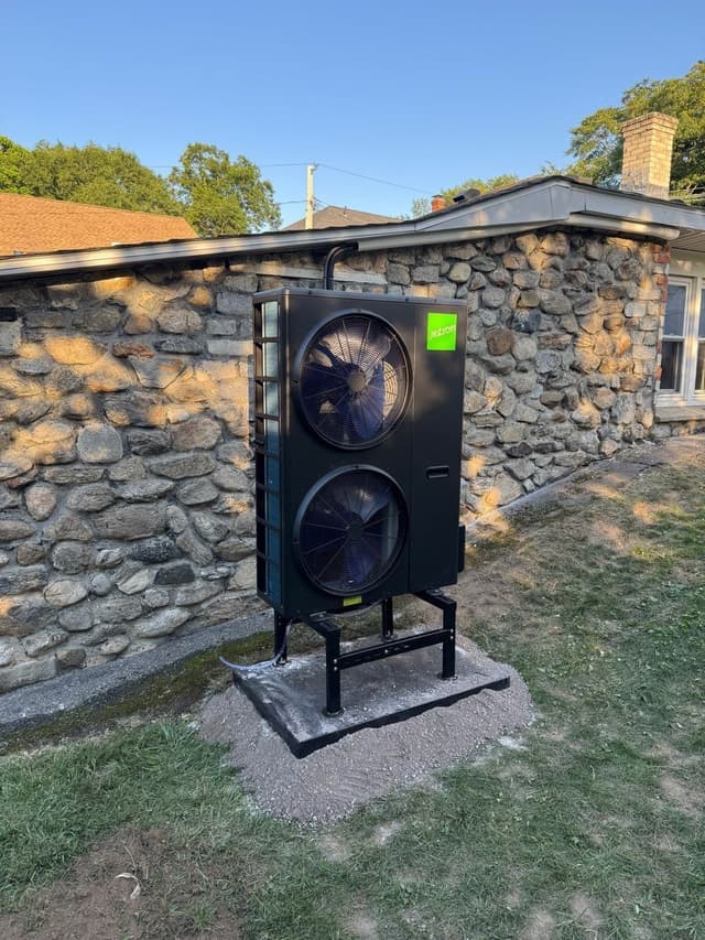 Black Jetson Air heat pump on a metal stand against the fieldstone exterior wall of a single-story home in Shrewsbury, MA, installation complete.