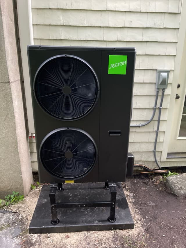 A large black Jetson Air 4-ton heat pump on a metal stand between house siding and a concrete wall in South Salem, NY, with gray conduit and copper linesets neatly run.