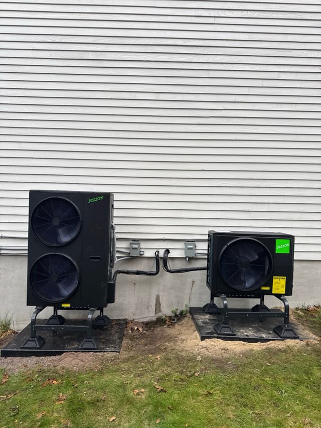 Two Jetson Air heat pump condensers installed side by side along the foundation of a white-sided home in Stoneham, MA, with line sets and disconnect boxes mounted on the wall between them.