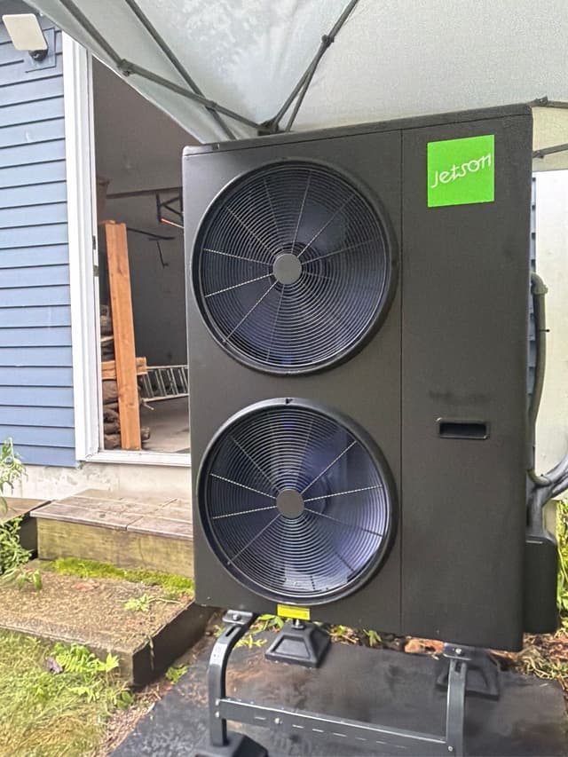 Black Jetson Air 4-ton central ducted heat pump installed beside a blue-sided house in Stow, MA, with a pop-up canopy tent visible overhead.