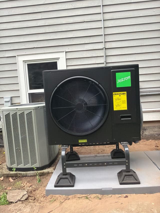 Jetson Air 3-ton central ducted heat pump mounted on an elevated stand against gray vinyl siding at a Sudbury home, with a smaller older condenser unit visible to the left.
