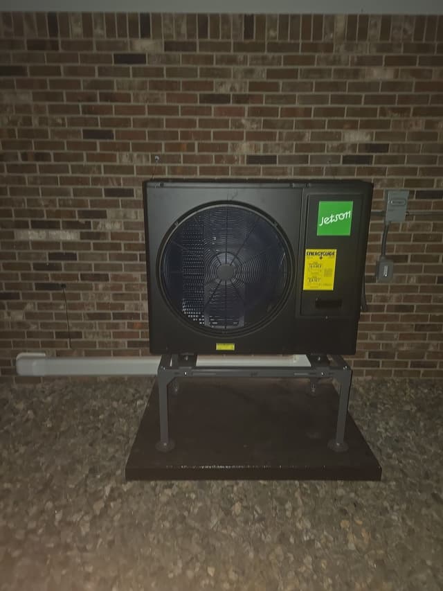 Jetson Air 3-ton central ducted heat pump mounted against a red brick wall in Thornton, with a grey disconnect box to the right, photographed at night.
