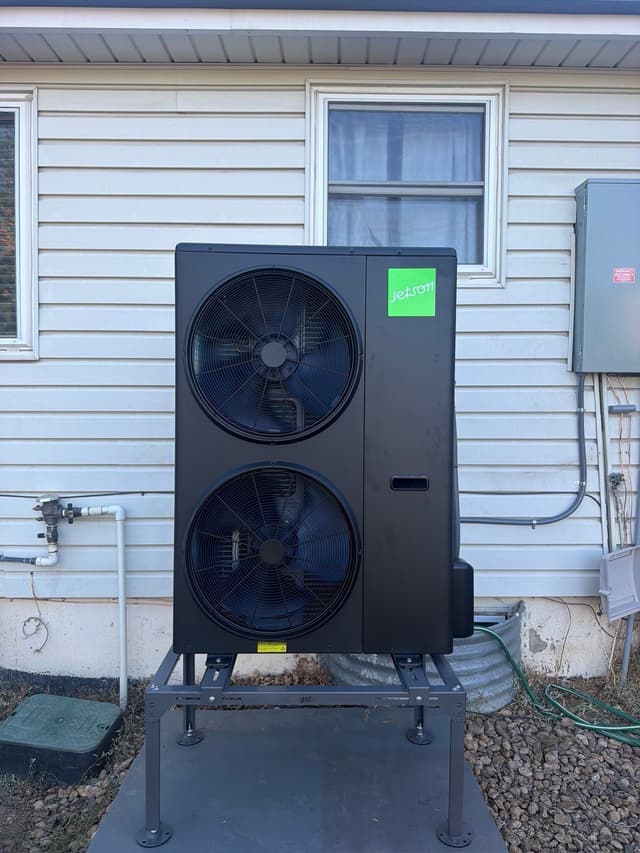 A large black Jetson Air 4-ton heat pump installed along the side of a white vinyl-sided house in Thornton, with electrical disconnect box and utility piping visible on the wall.