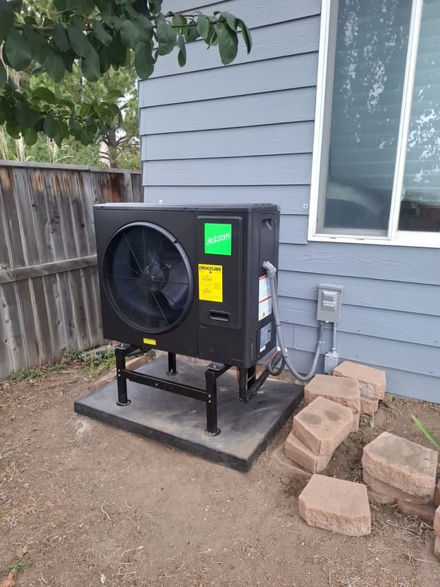Black Jetson Air 3-ton central ducted heat pump condenser installed along the side of a blue-gray lap-siding house in Thornton, with electrical conduit running to a disconnect box and a wooden fence in the background.