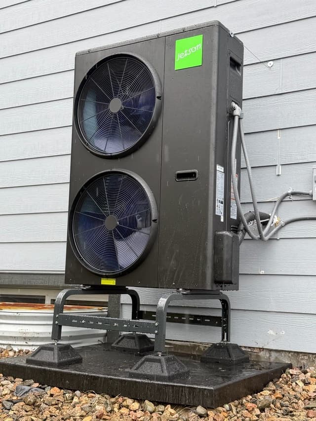 Jetson Air 5-ton dual-fan outdoor heat pump condenser mounted on a black riser stand over a gravel pad against a grey lap-siding house in Thornton, CO.