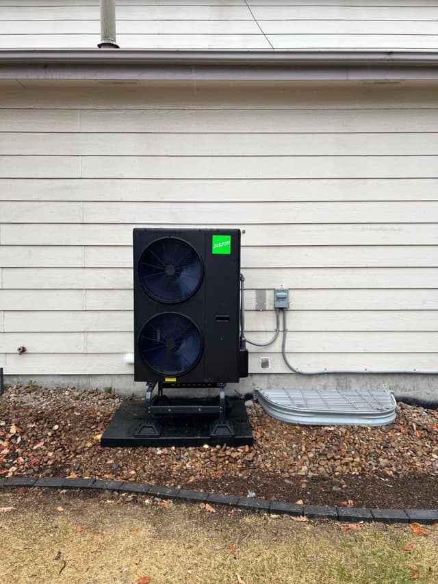 Black Jetson Air 4-ton heat pump on an elevated stand in a gravel bed against white vinyl siding, with electrical disconnect box and conduit mounted on the wall, in Thornton, CO.