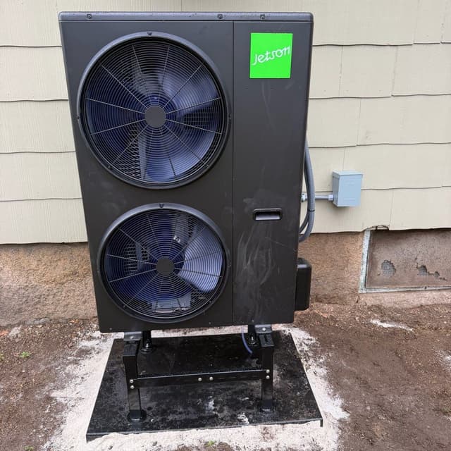 Jetson Air 4-ton heat pump mounted on a black metal stand on a concrete pad along the side of a house in Uxbridge, MA, with conduit running to a gray electrical disconnect box on the wall.