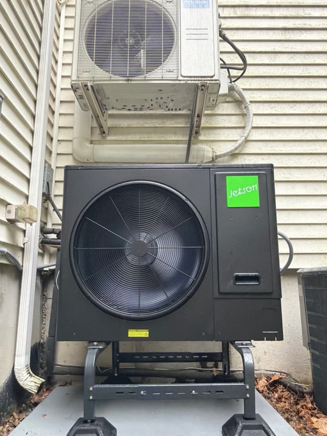 Jetson Air 3-ton central ducted heat pump mounted on a metal wall-bracket stand against light vinyl siding of a house in Uxbridge, MA.