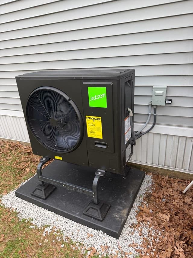 Black Jetson Air 3-ton heat pump condenser on a black equipment stand over a gravel and rubber pad base, mounted against gray vinyl siding with conduit running to a gray disconnect box, Webster, MA.