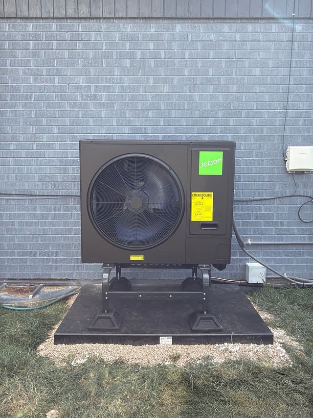 Jetson Air 3-ton heat pump mounted on an elevated stand against a grey brick building in Westminster, with conduit and disconnect box visible on the wall.