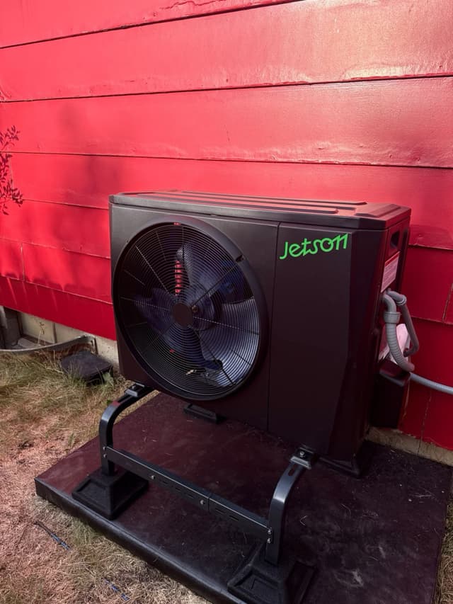 Black Jetson Air heat pump condenser on a metal equipment stand against a bright red horizontal-plank wall in Wheat Ridge, CO.