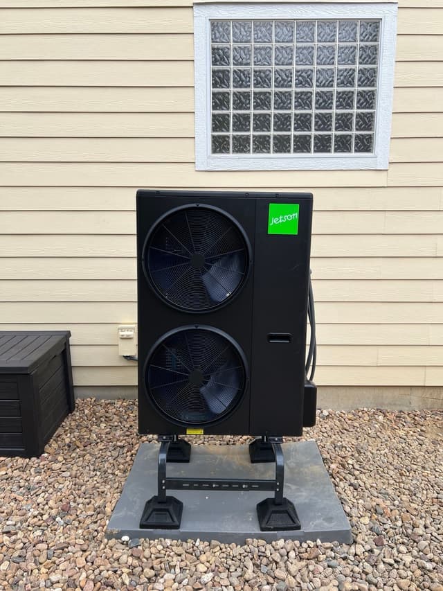 Black Jetson Air 5-ton central ducted heat pump unit installed against beige vinyl siding with a glass-block window above, in Windsor, CO.